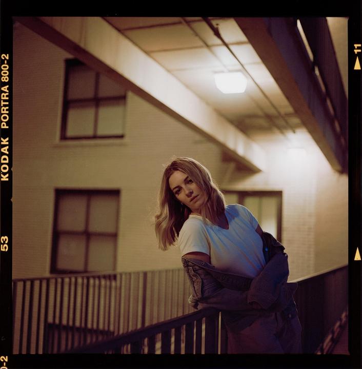 A woman leans on a railing at night with a building and overhead lights in the background.