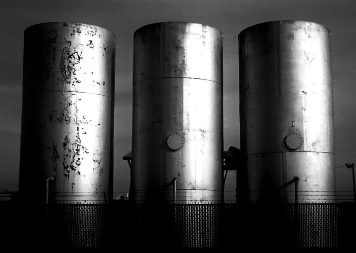 Three large industrial silos stand against a dark sky, with a fence visible at their base.