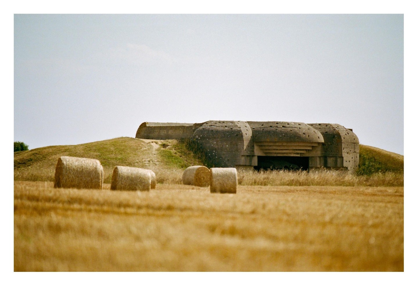 A concrete bunker is partially hidden by a grassy hill and several hay bales in a field.