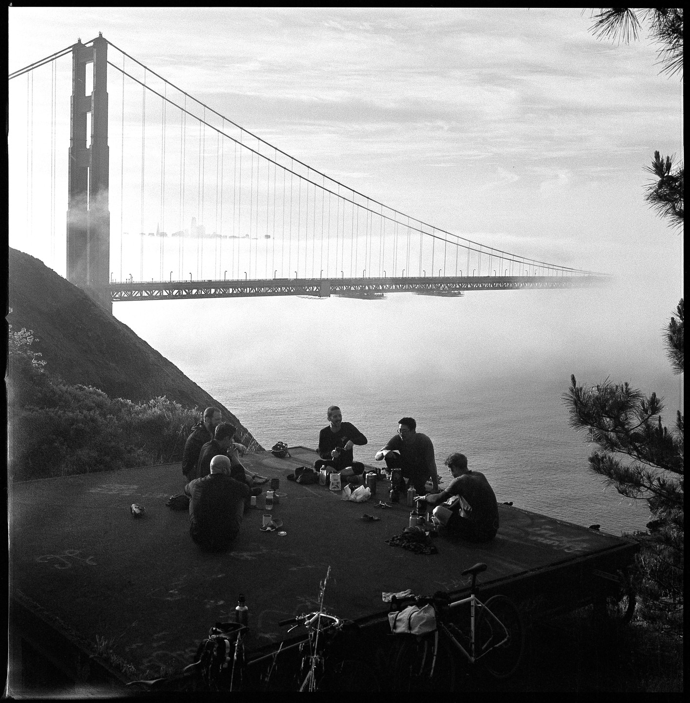 A group of men sit on a cliff overlooking the Golden Gate Bridge, which is partially obscured by fog.