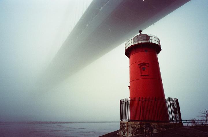 A red lighthouse stands on a rocky shore under a bridge on a hazy day.
