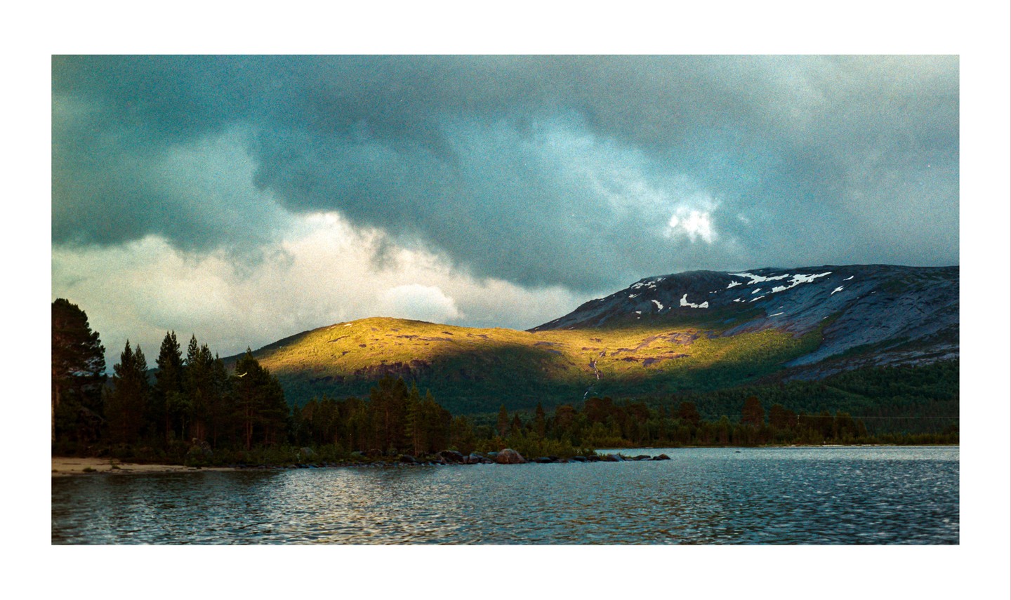 A lake with trees on its shore is overlooked by mountains, one of which is illuminated by a cloud break.