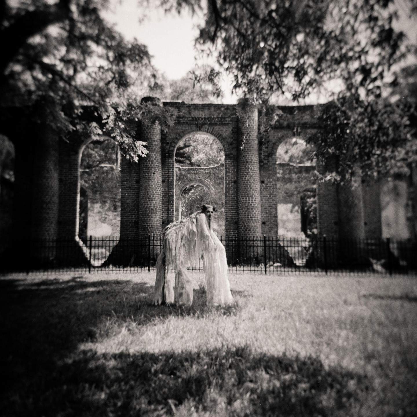A person in a costume resembling a shaggy animal stands in a grassy area in front of brick ruins with arched openings.