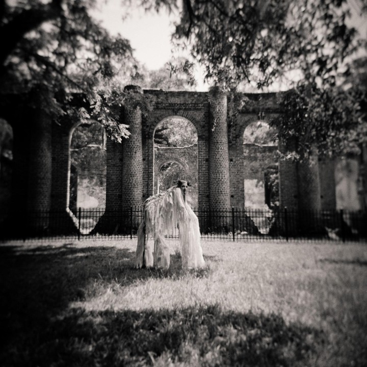 A person in a costume resembling a shaggy animal stands in a grassy area in front of brick ruins with arched openings.