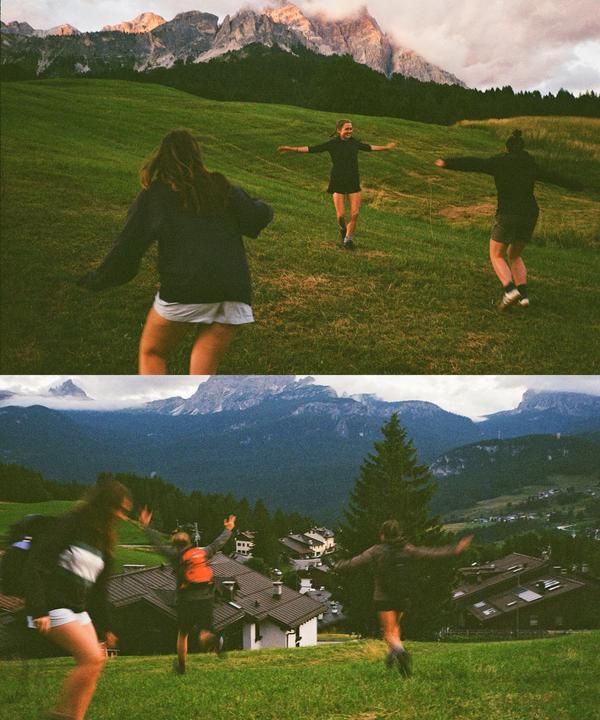 Three women run down a grassy hill with mountains in the background.