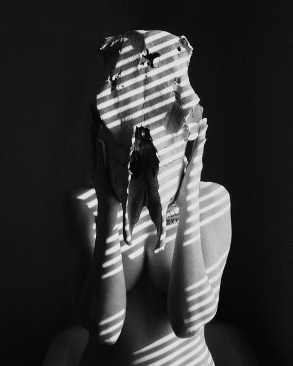A person with a skull mask covers their face with their hands, illuminated by striped shadows.