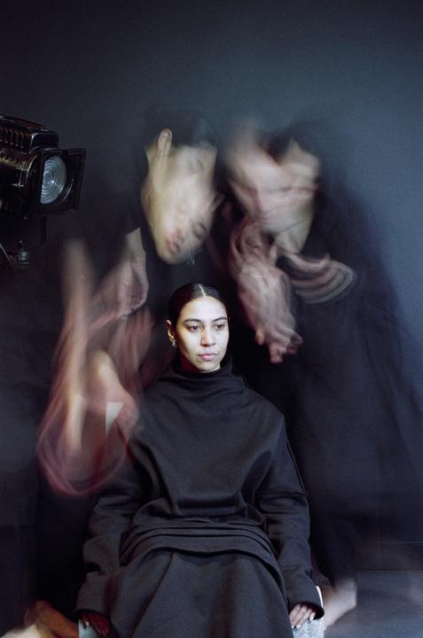 A woman sits in a dark room with blurred figures and a spotlight in the background.