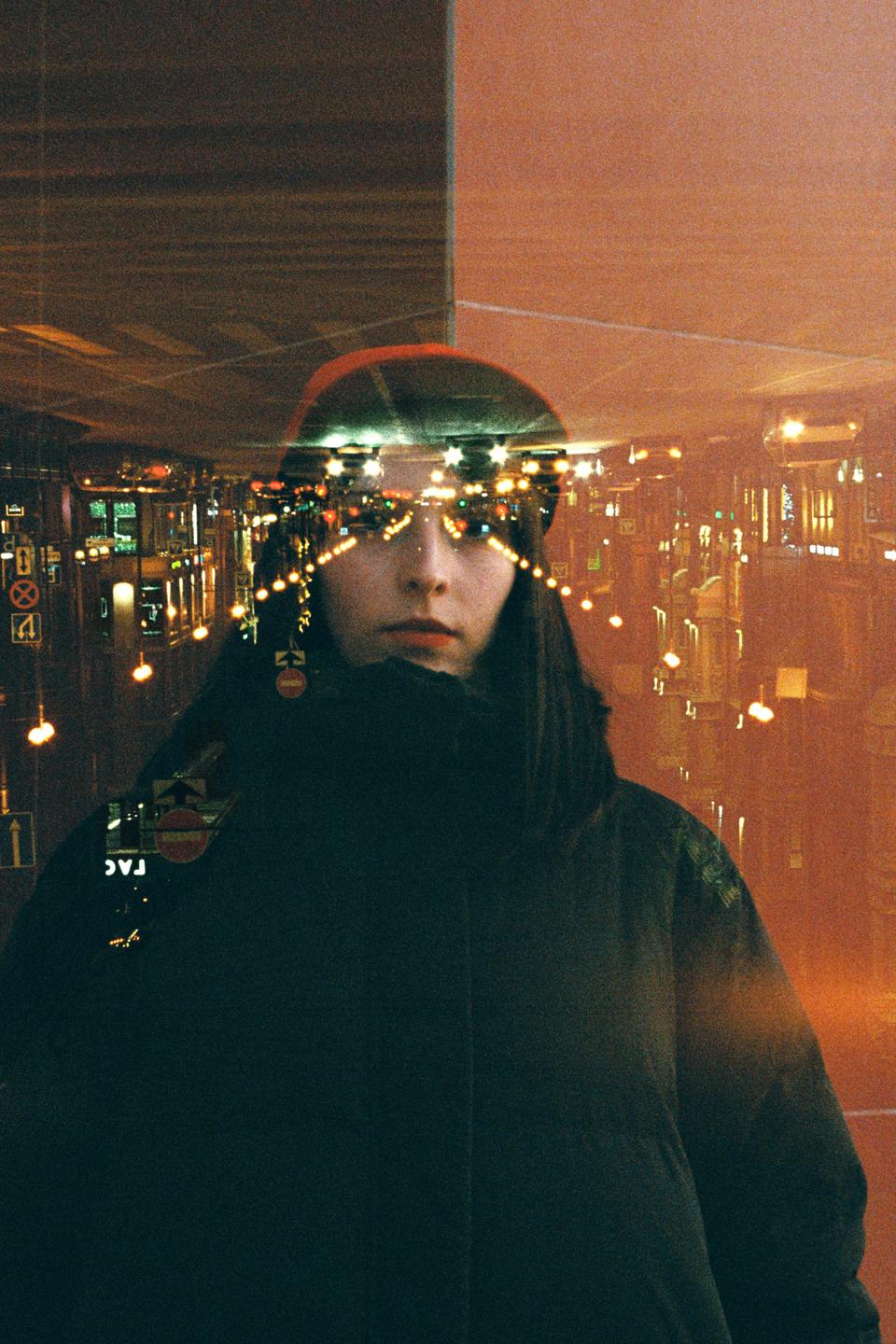 A woman with long hair and a dark jacket is visible through a double exposure of city lights at night.