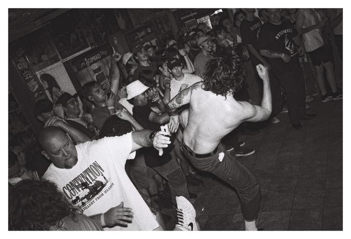 A shirtless man with long hair dances in a crowded room with other men.