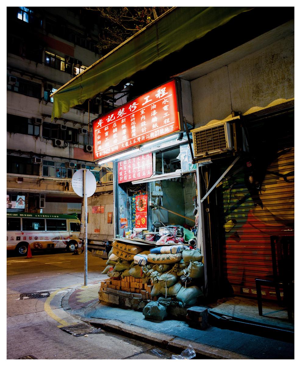 A street corner in Hong Kong is illuminated by a red neon sign above a shop with sacks and bricks piled outside.