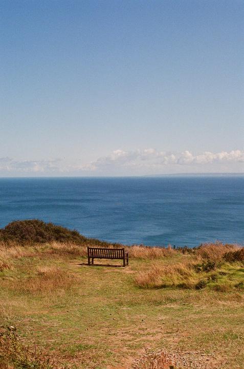 A wooden bench sits on a grassy cliff overlooking a vast blue ocean under a clear sky.