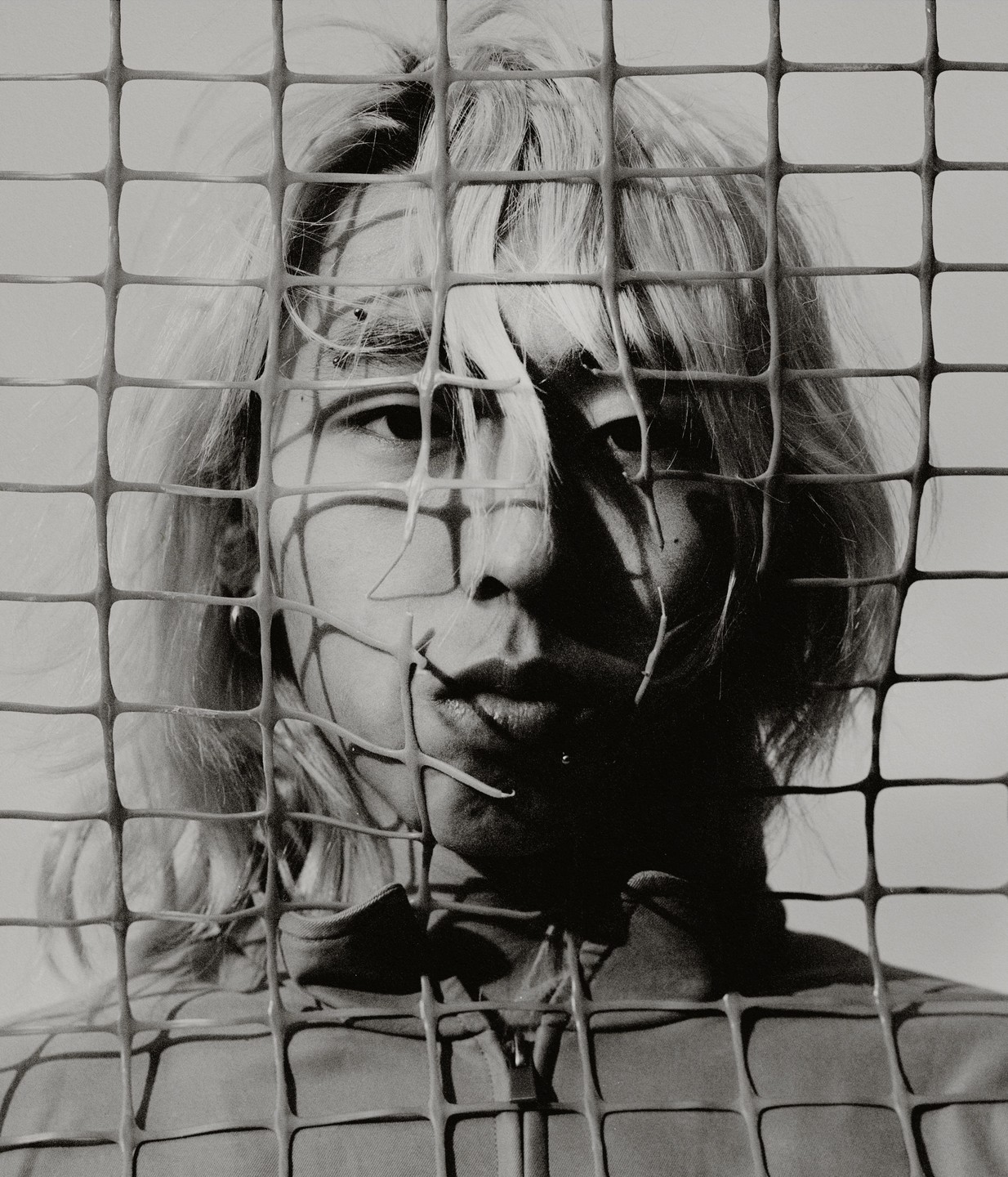A person with light hair looks through a grid fence, with shadows cast across their face.