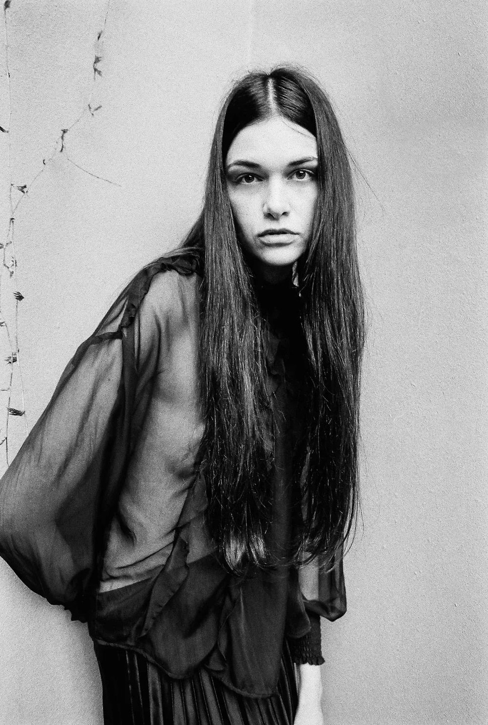 A woman with long hair wears a sheer top and looks directly at the viewer.
