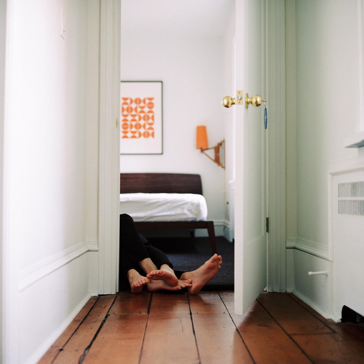 Two pairs of feet are visible on a dark rug in a bedroom, seen from a hallway with a wooden floor.