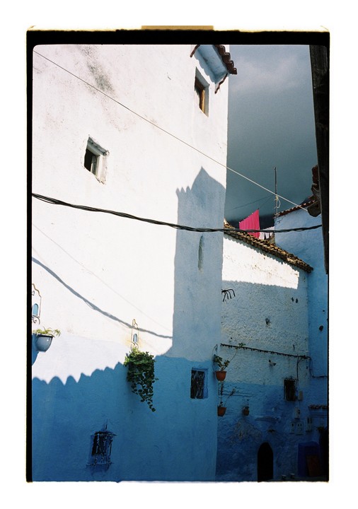 Sunlight casts shadows on blue and white buildings with potted plants and laundry hanging in Chefchaouen.