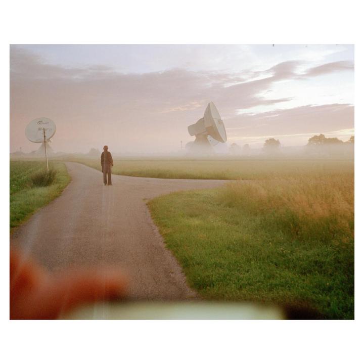 A person stands on a road in a rural landscape with a large satellite dish in the distance under a hazy sky.