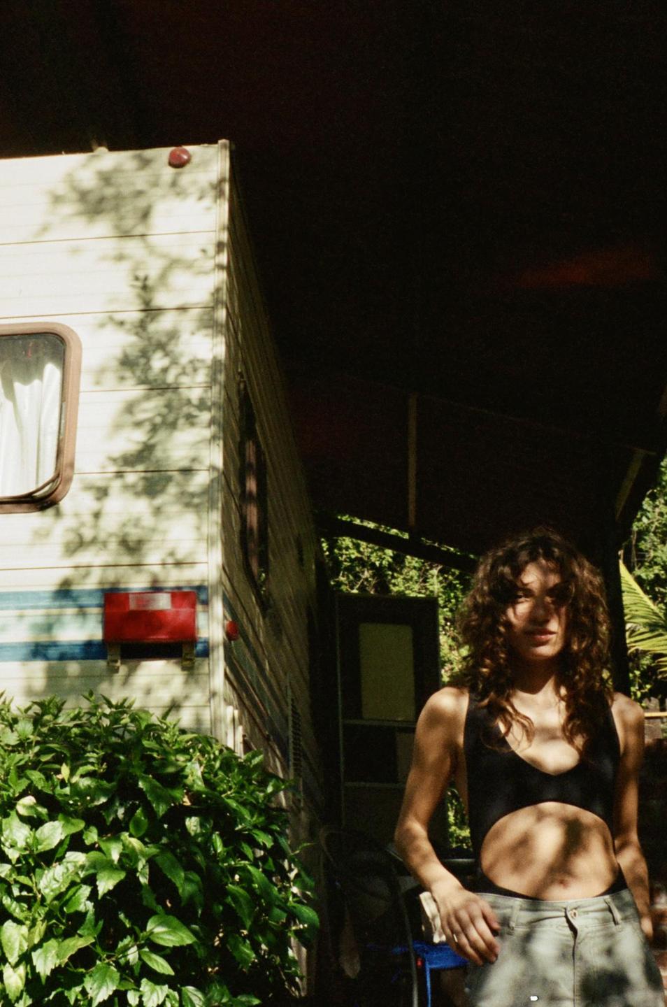 A woman with curly hair stands next to a camper van, with dappled sunlight on her body and the side of the vehicle.