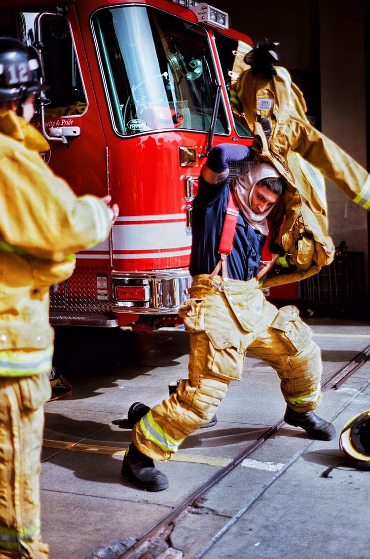 A firefighter puts on his gear next to a fire truck while another firefighter stands nearby.