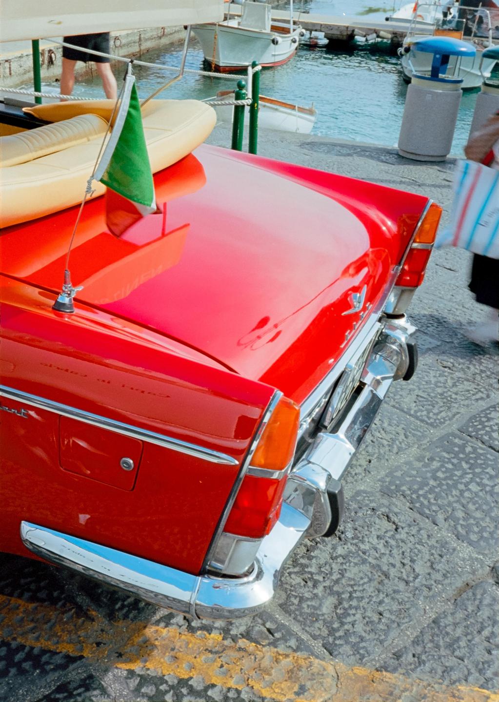 A red vintage car with a small Italian flag is parked on a stone street next to a body of water with boats.