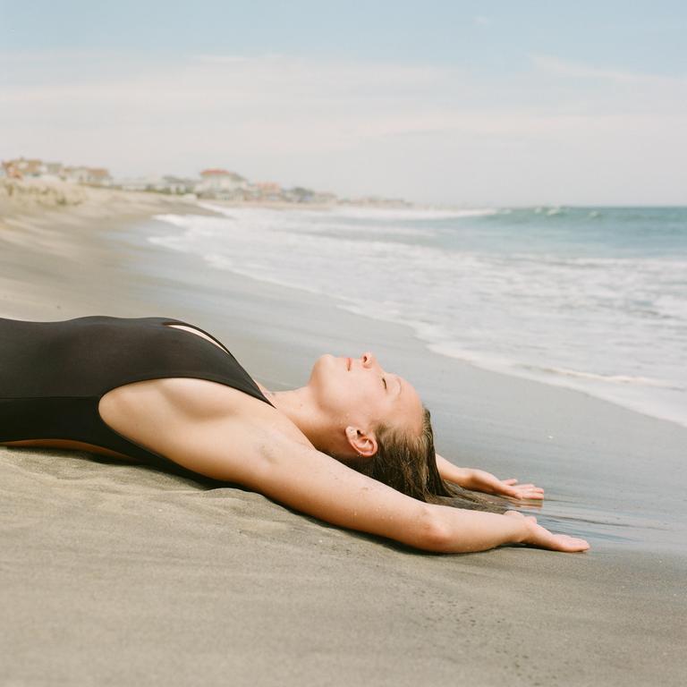 A woman in a black swimsuit lies on a sandy beach with the ocean in the background.