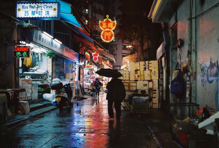 A person holding an umbrella walks down a wet street at night past shops with neon signs and stacked boxes.