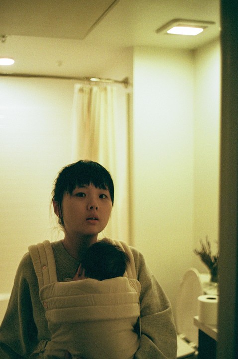 A woman with a baby in a carrier stands in a bathroom.