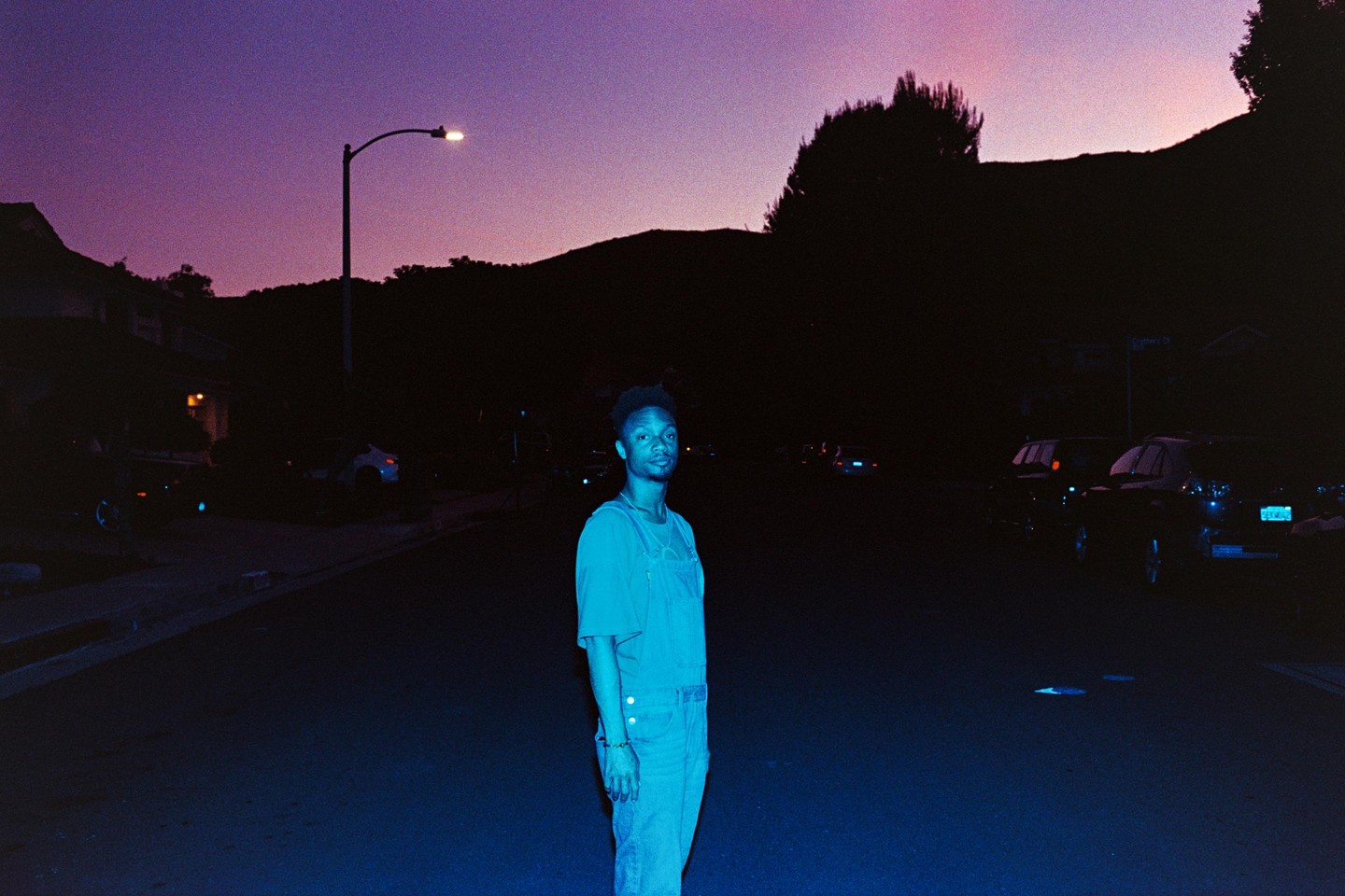 A man stands in the middle of a street at dusk, illuminated by a street light.