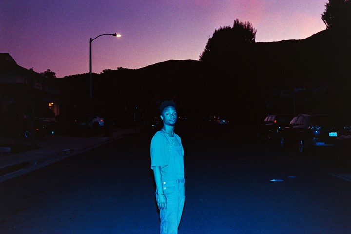 A man stands in the middle of a street at dusk, illuminated by a street light.