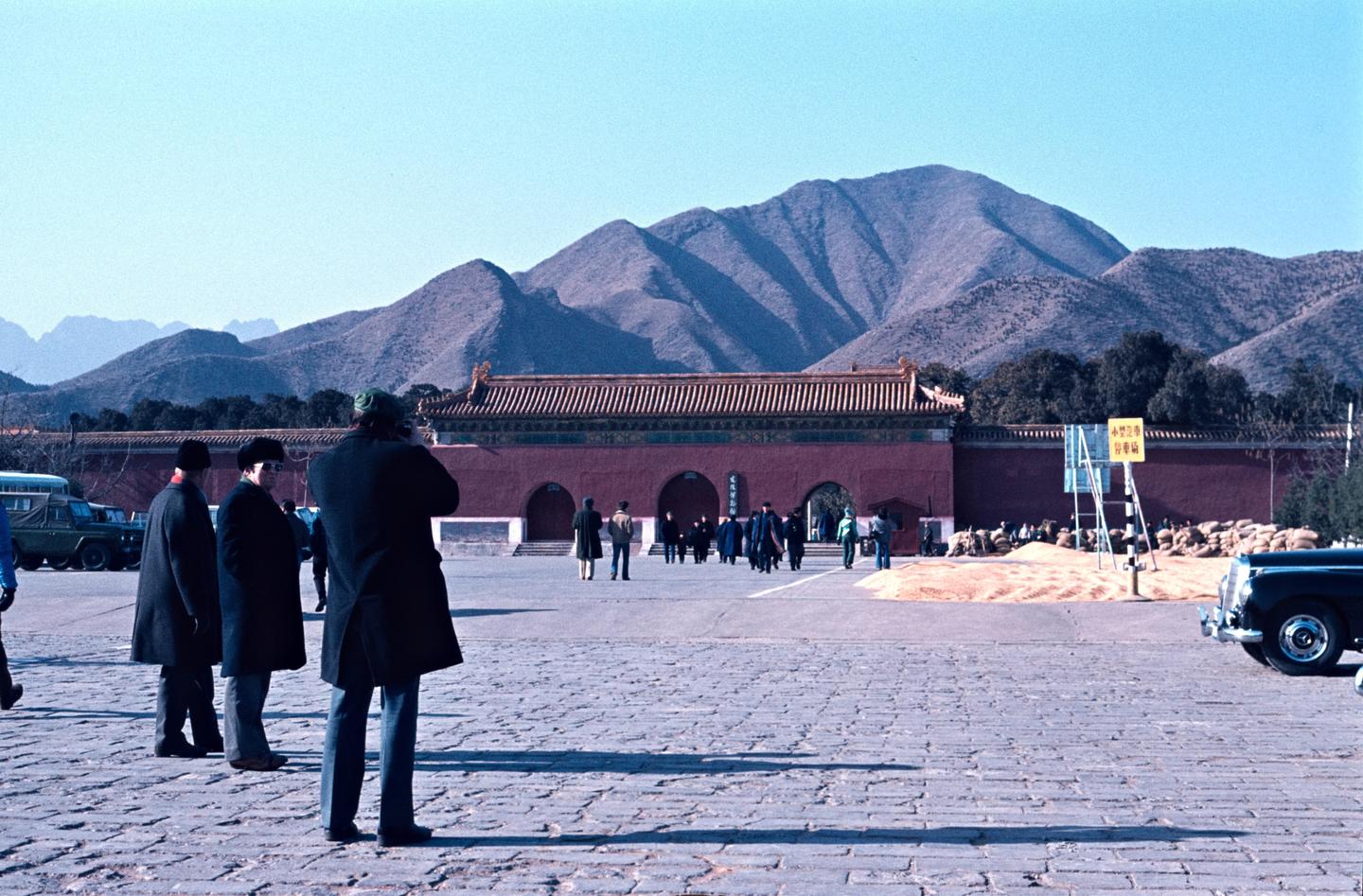 Several people stand on a paved area in front of a red building with a tiled roof, with mountains in the background.