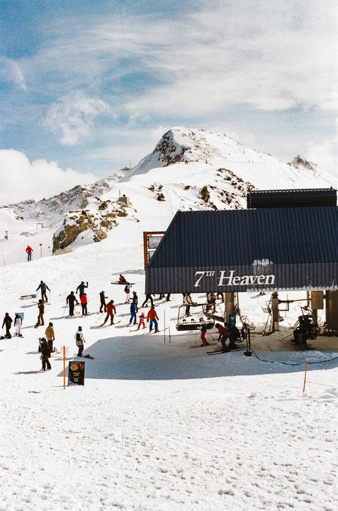 A group of skiers and snowboarders gather at the base of a ski lift named "7th Heaven" on a snowy mountain.