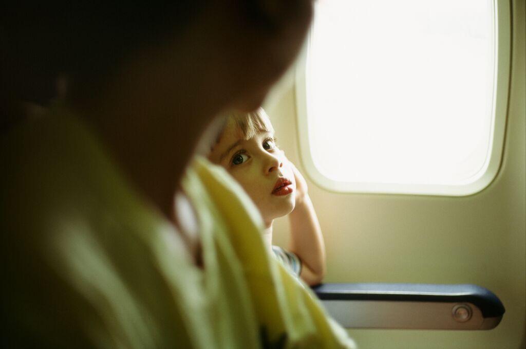 A child with light hair looks over the shoulder of another person, with a bright window visible to the right.