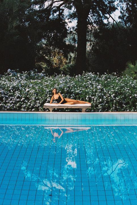 A woman in a black swimsuit is lying on a lounge chair next to a swimming pool, with a hedge of white flowers and trees in the background.