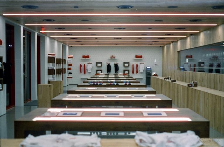 A clothing store interior features multiple display tables with illuminated strips and shelves of folded garments.