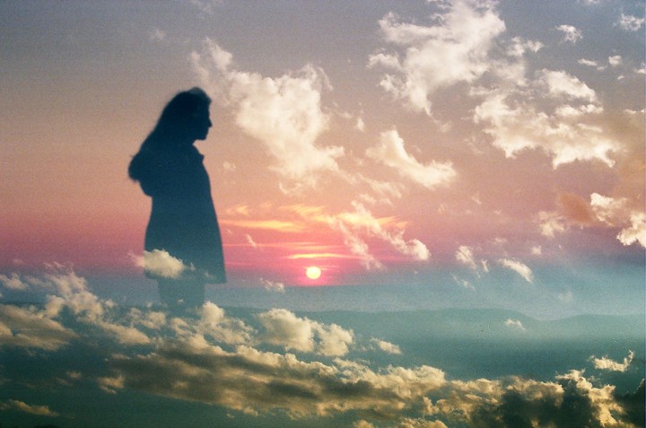 A silhouette of a woman is superimposed over a sunset sky with clouds and distant mountains.