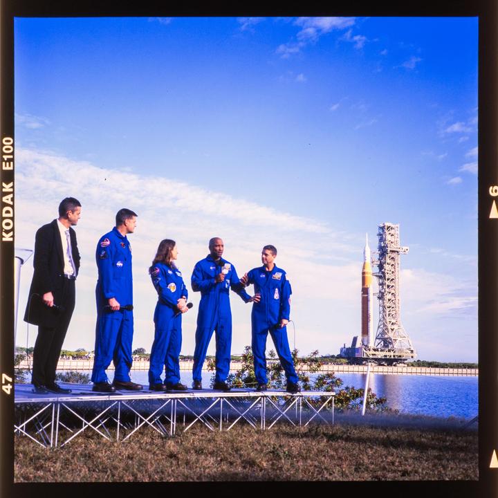 Five people stand on a stage with a rocket and launch tower in the background.