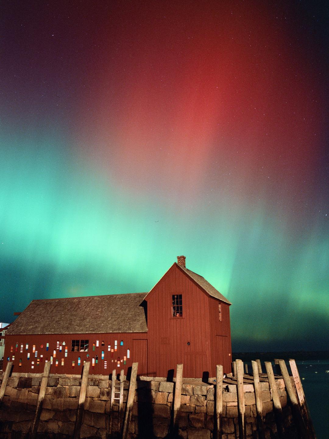 A red wooden building with a stone foundation stands on a pier under a night sky with aurora borealis.