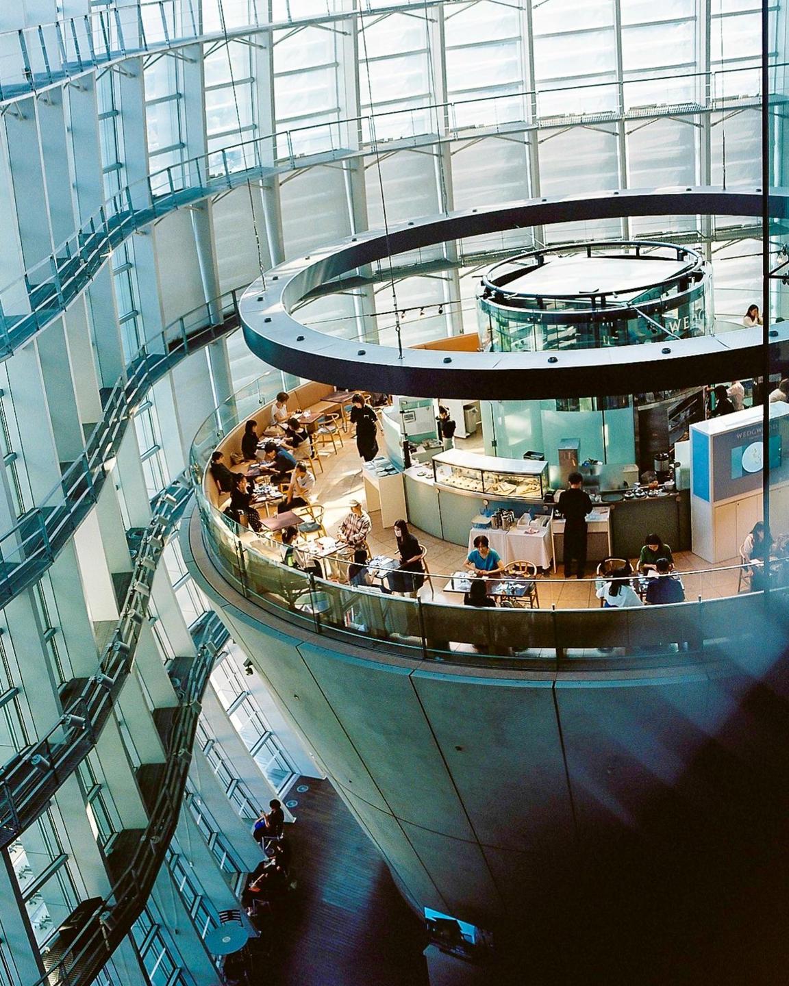 A cafe with people sitting at tables is visible from above, surrounded by a modern building with many windows.