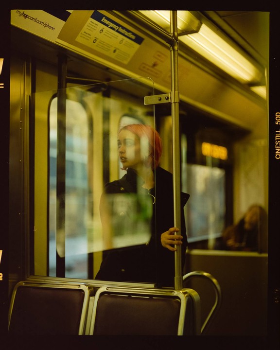 A woman with pink hair looks out a window on a train.