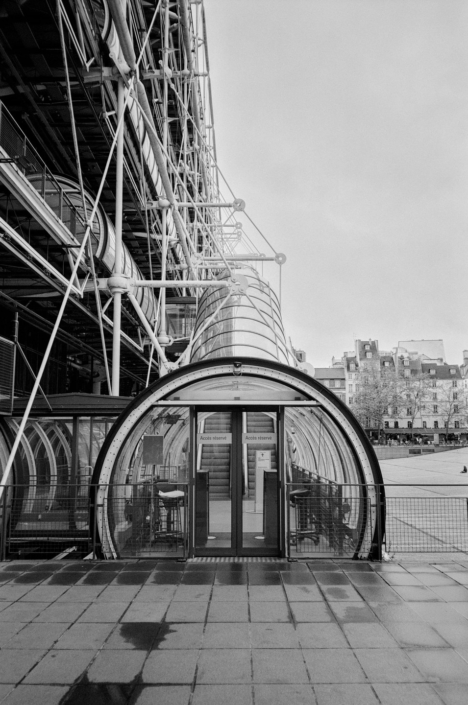 A black and white photograph shows the exterior of a modern building with an arched glass entrance and exposed structural elements.