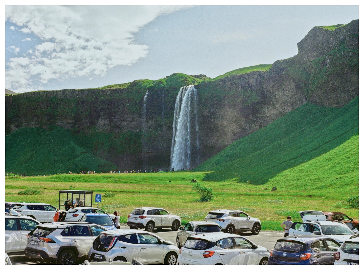 A parking lot full of cars sits in front of a large waterfall cascading down a green cliff.
