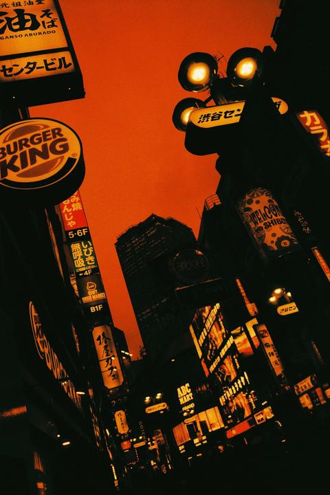 A low-angle view captures a vibrant cityscape at night, dominated by illuminated signs and buildings against an orange sky.