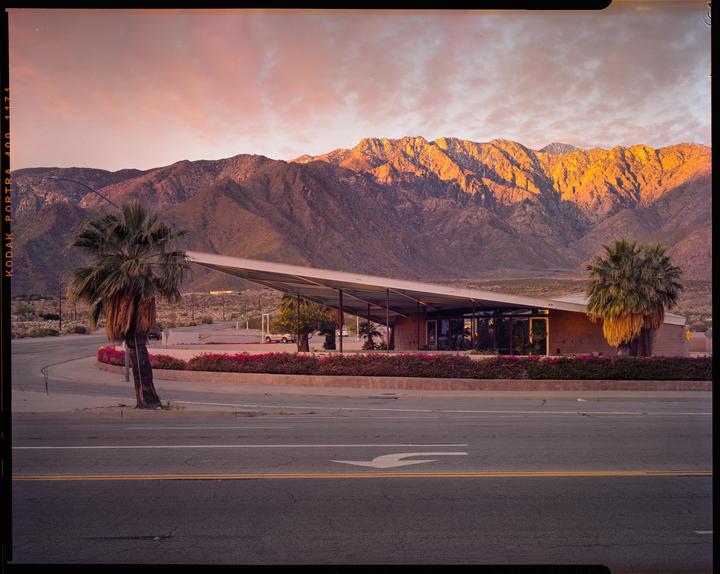 A modern building with a large angled roof stands in front of a mountain range under a colorful sky.