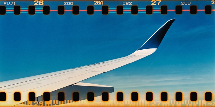 An airplane wing is visible against a clear blue sky, with film sprockets visible at the top and bottom of the image.