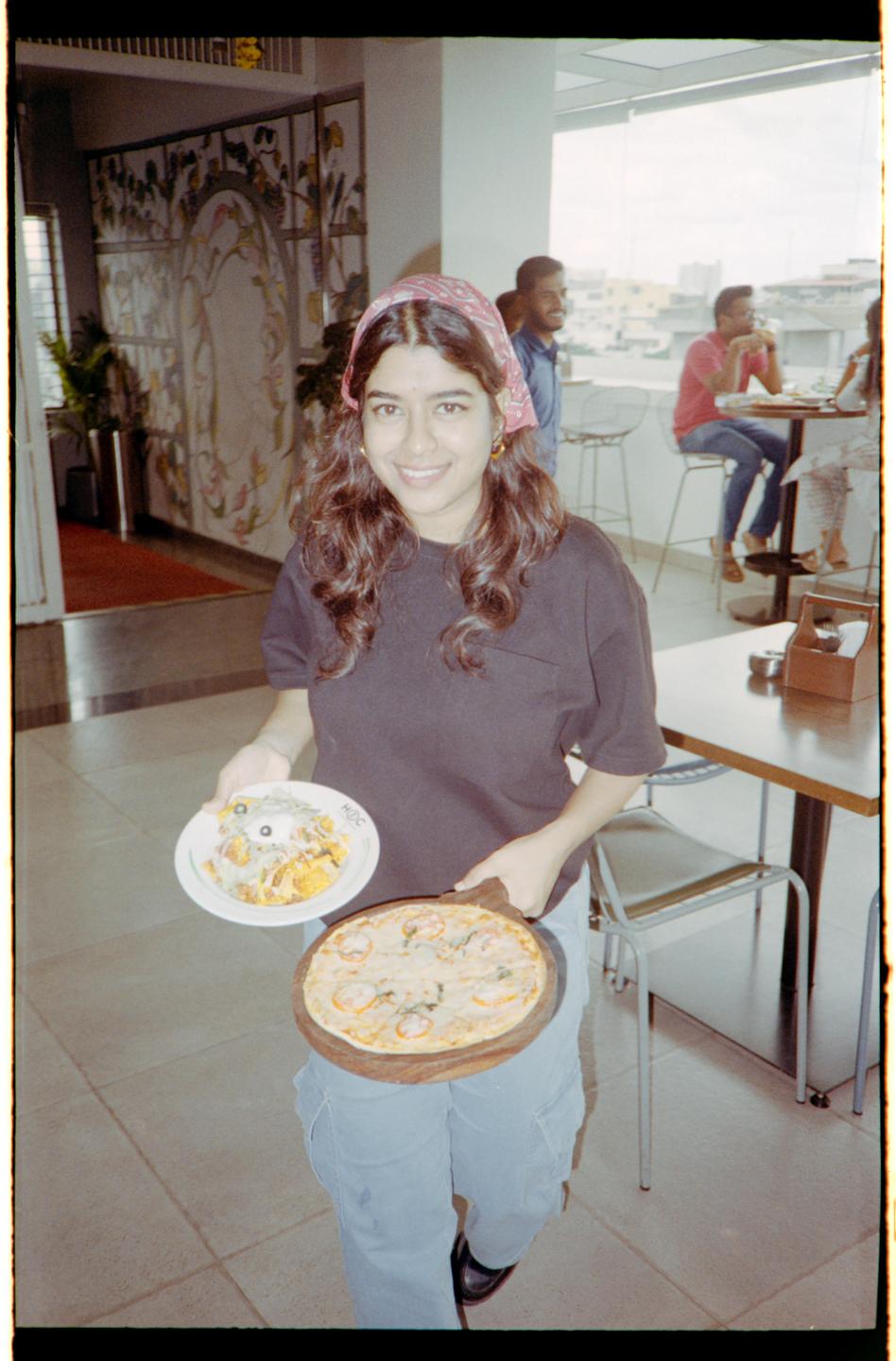 A woman smiles while holding a pizza and a plate of food in a restaurant.