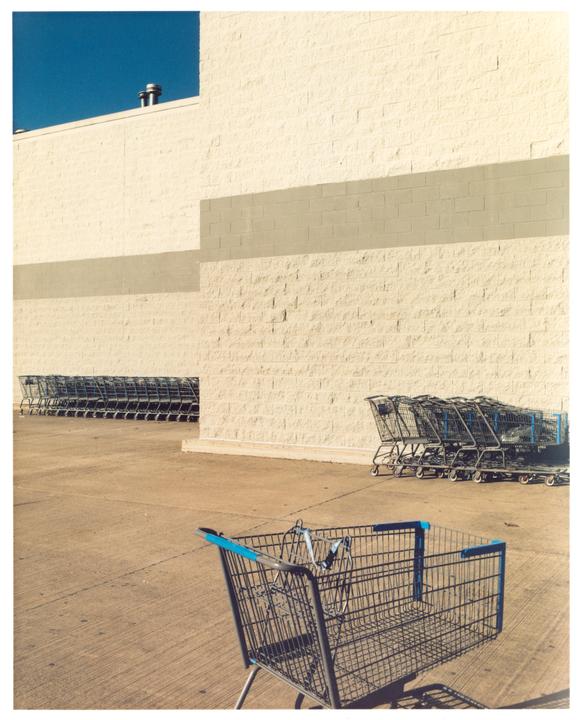 A single shopping cart sits in a parking lot with a large building and rows of other shopping carts in the background.