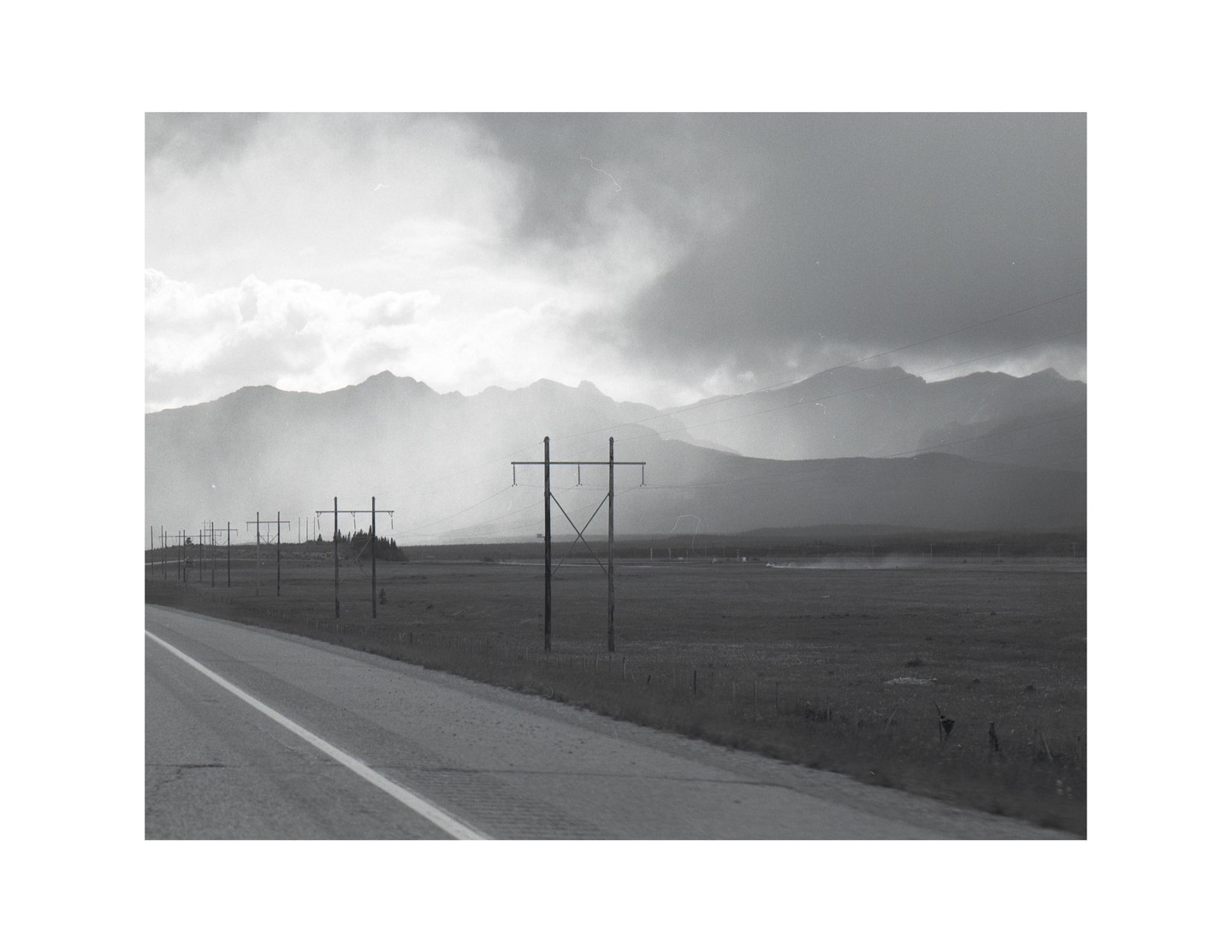 A long road stretches into the distance, lined with telephone poles and mountains under a cloudy sky.