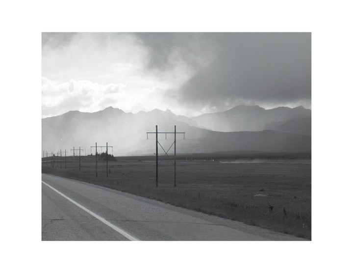A long road stretches into the distance, lined with telephone poles and mountains under a cloudy sky.