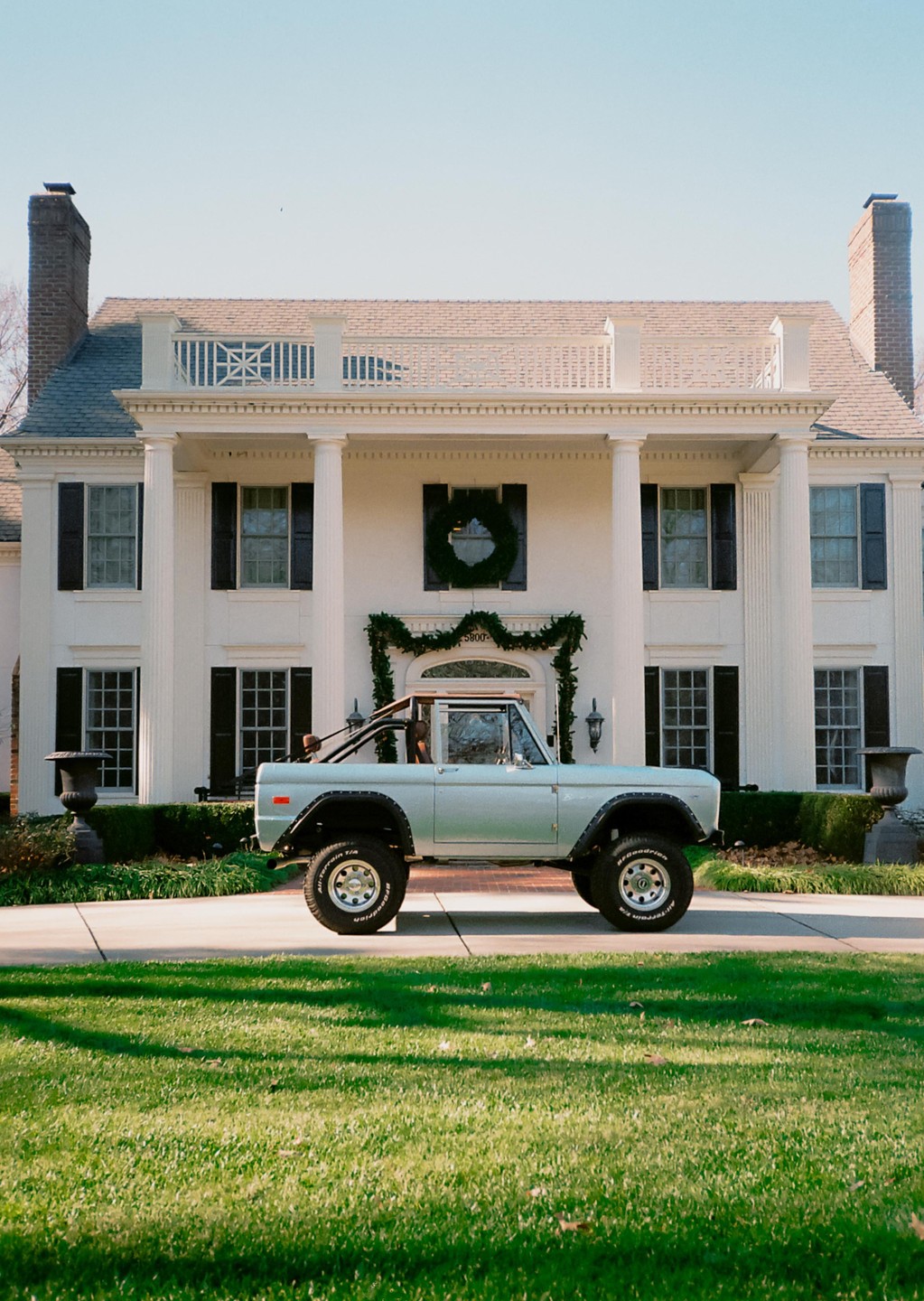 A light blue Ford Bronco is parked in front of a large white house with columns and a decorated entrance.