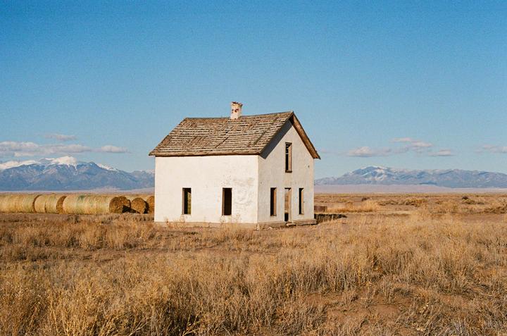 An abandoned house stands in a dry field with mountains in the distance.