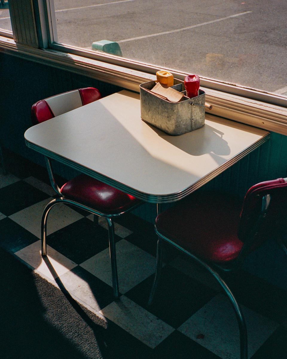 A diner booth with a table and two chairs sits next to a window, with a condiment caddy on the table.